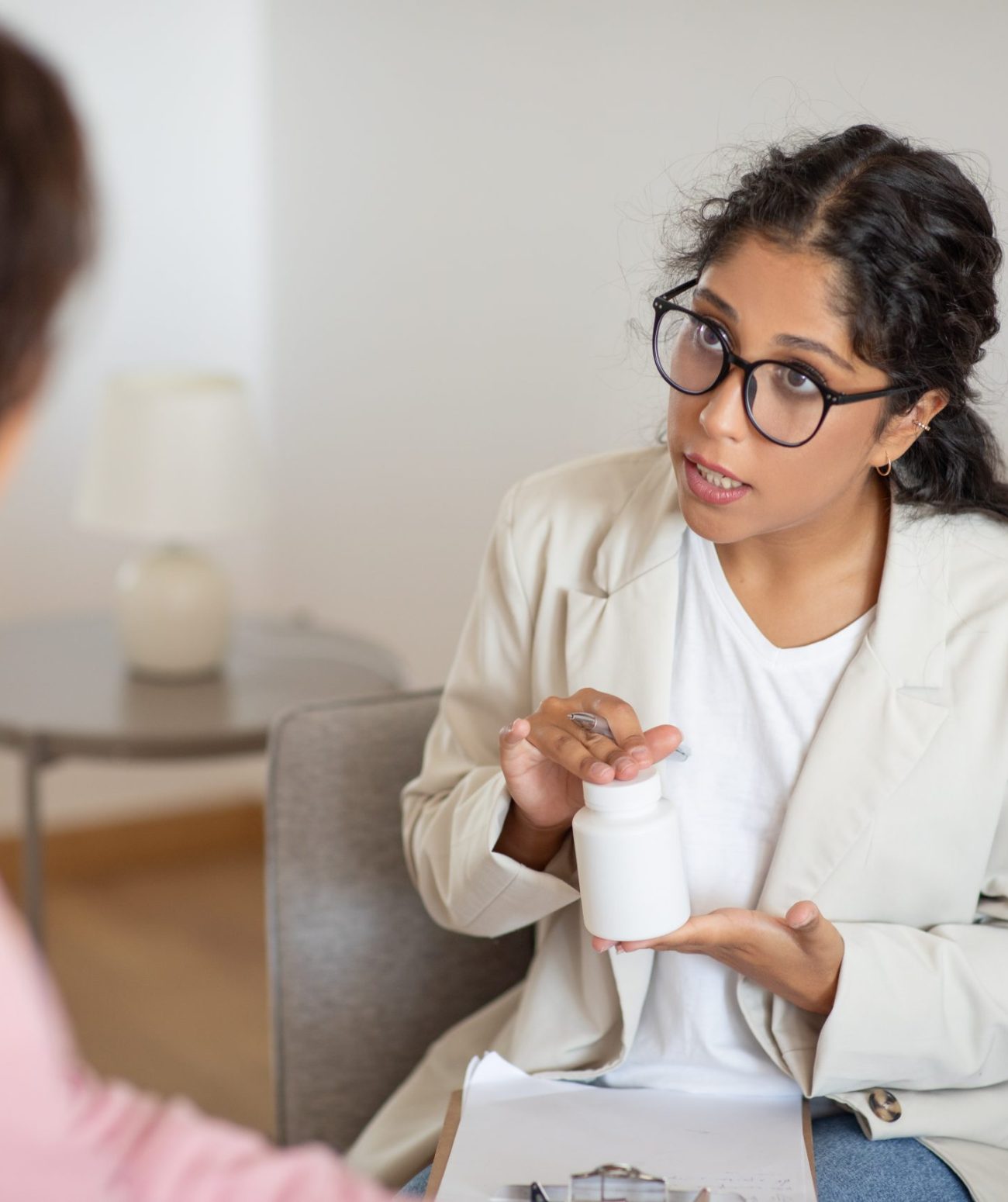 A woman in a white coat is seated across from a patient, engaging in a detailed explanation about a medication. She holds a pill bottle in her hand and gestures to signify the directions for use