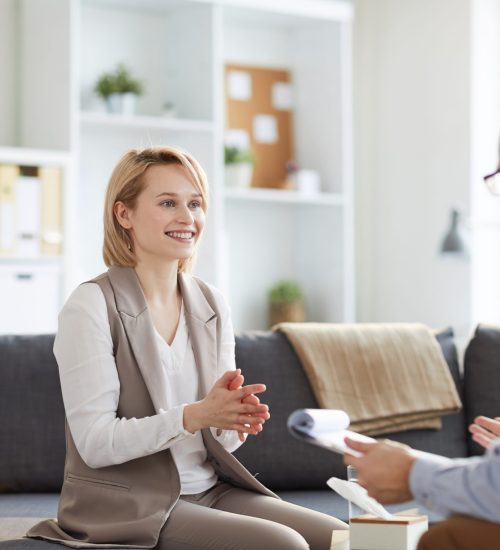 Young blonde smiling woman sitting on couch and looking at counselor during discussion of ways of solving problem