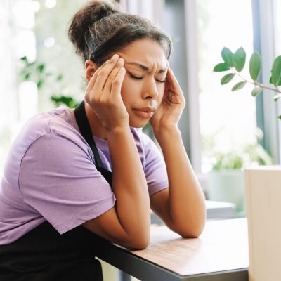 Young waitress holds her head in pain, showing stress and exhaustion at work in a cafe. The image captures the reality of overworked service industry employees